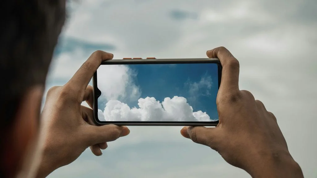 Person holding a smartphone horizontally while photographing white clouds against a blue sky.