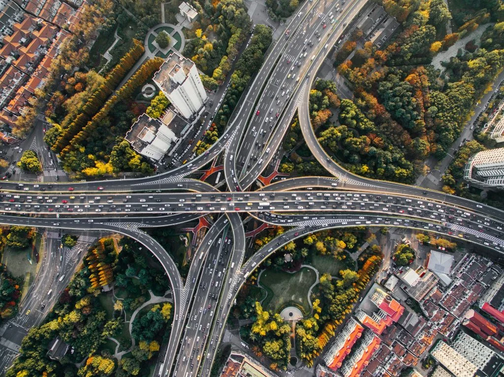 Aerial view of a large multi-level highway interchange with heavy traffic flowing in multiple directions, surrounded by trees, parks and city buildings.