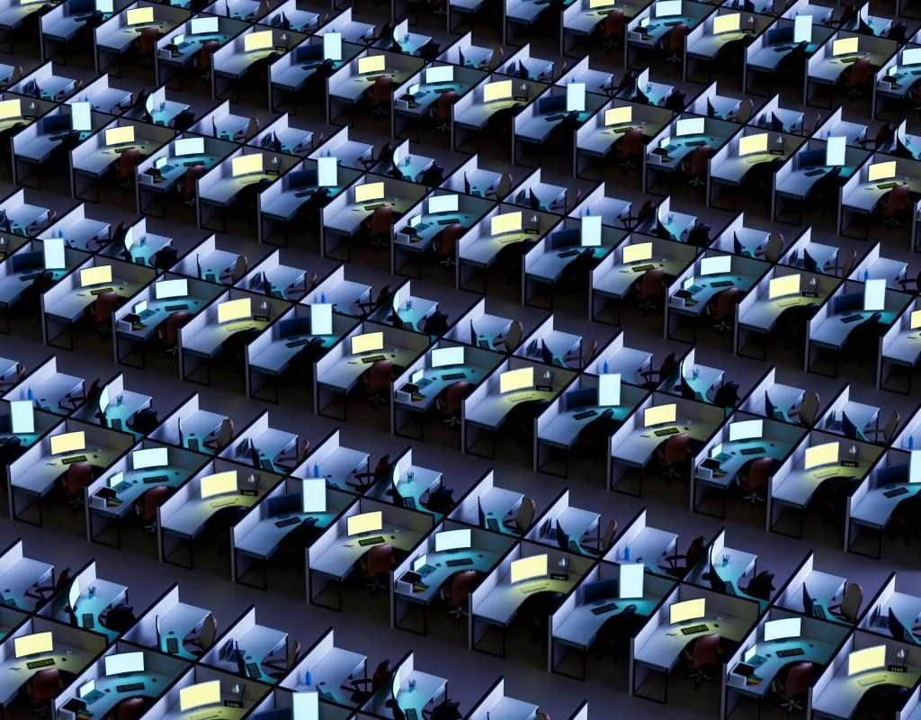 Aerial view of a vast, dimly lit office floor filled with rows of identical cubicles, each containing a desk, chair, and glowing computer monitor stretching into the distance
