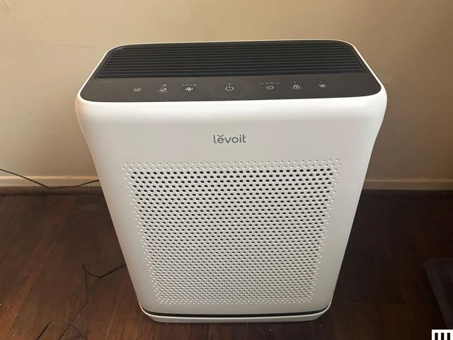 Front of white rectangular Levoit Vital 200S-P Air Purifier. Sits on wood floor with white wall behind.