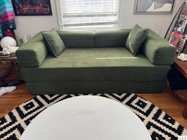 Mercer41 Convertible Sleeper Sofa, a long green rectangular sofa up against a wall with a window and framed artwork behind it. There's a black and white patterned circle rug and white circular table in front.