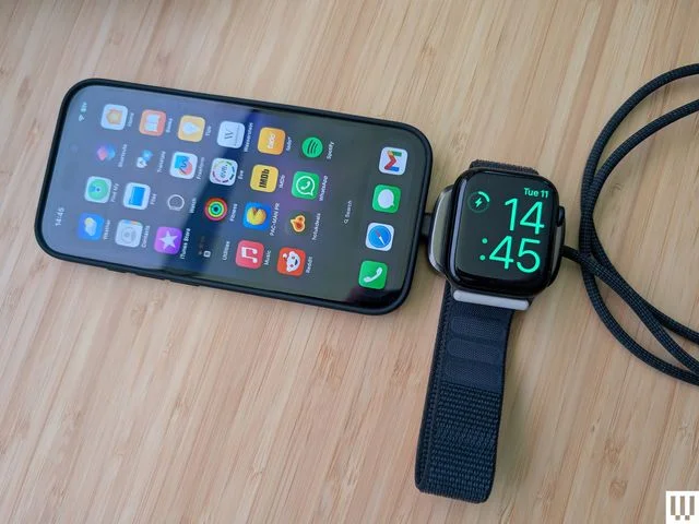 Smartphone with a charging pad plugged into it's bottom port. A smartwatch rests on the pad, a long black cord coming out the other end. 