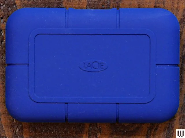 Overhead view of a LaCie Rugged SSD Pro5 external hard drive, a blue rounded rectangular device on a wooden surface