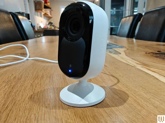 Oval shaped indoor security camera by Arlo with white base and black front, sitting on wooden surface